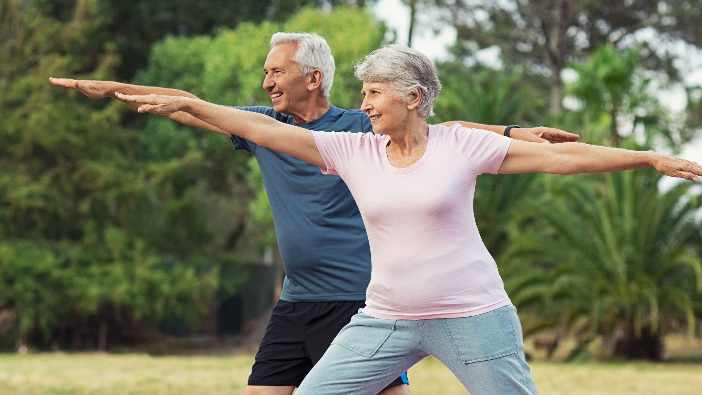 Stretching e declino cognitivo: una donna e un uomo anziani praticano la posizione del guerriero yoga al parco.