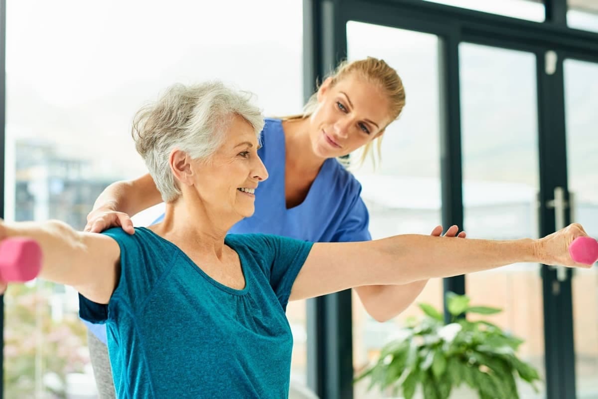 Donna anziana che esegue esercizi con pesi leggeri assistita da una fisioterapista, simbolo del ruolo della vitamina D su forza muscolare ed equilibrio