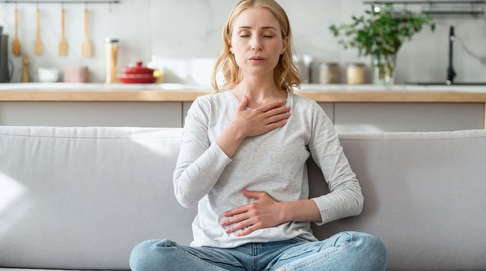 Donna seduta con occhi chiusi e mani su petto e addome mentre pratica respirazione consapevole, esercizio di mindfulness, in un ambiente domestico tranquillo
